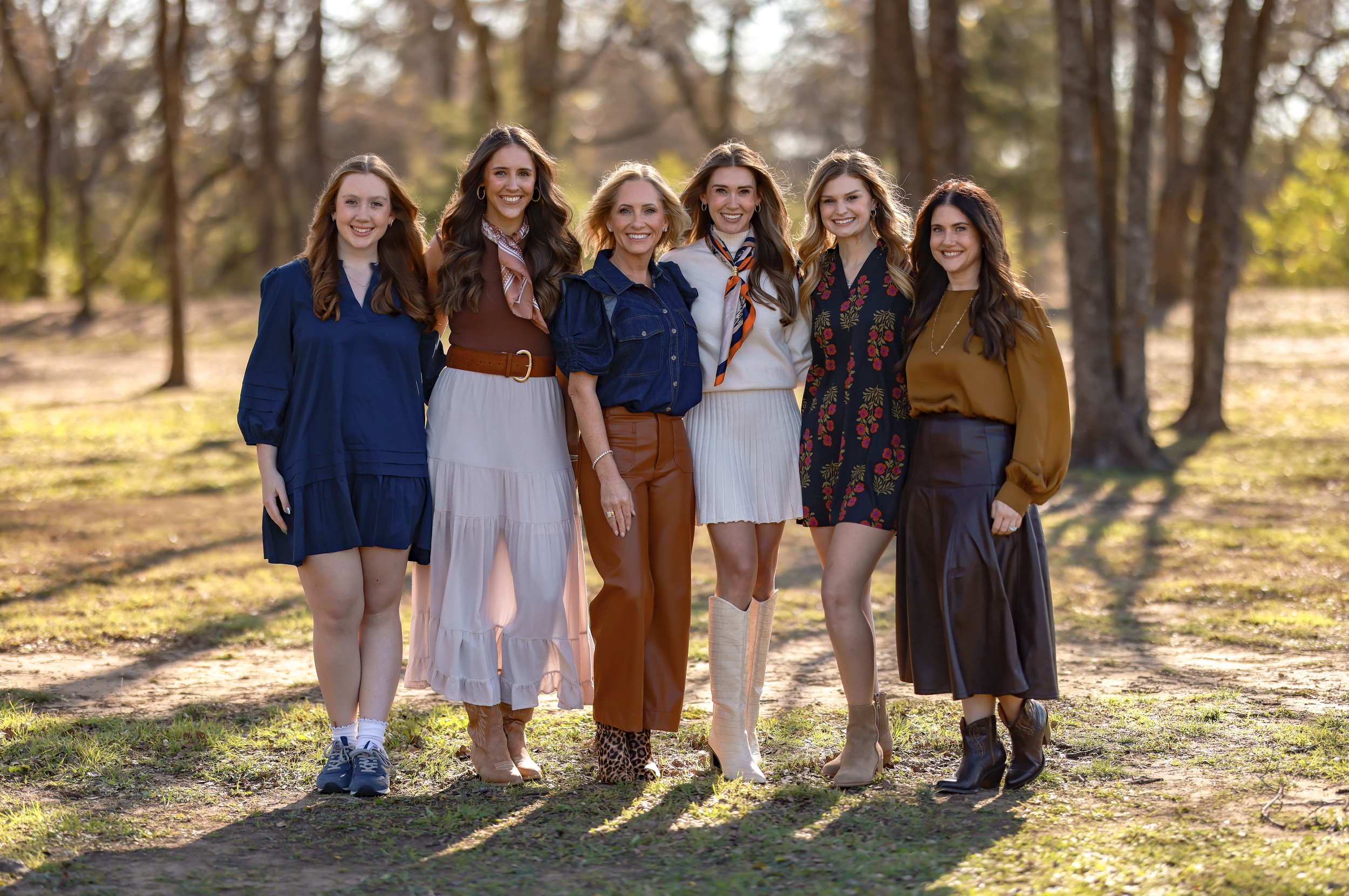 Group of six women standing outdoors in a wooded area, smiling at the camera, dressed in fall-inspired clothing.