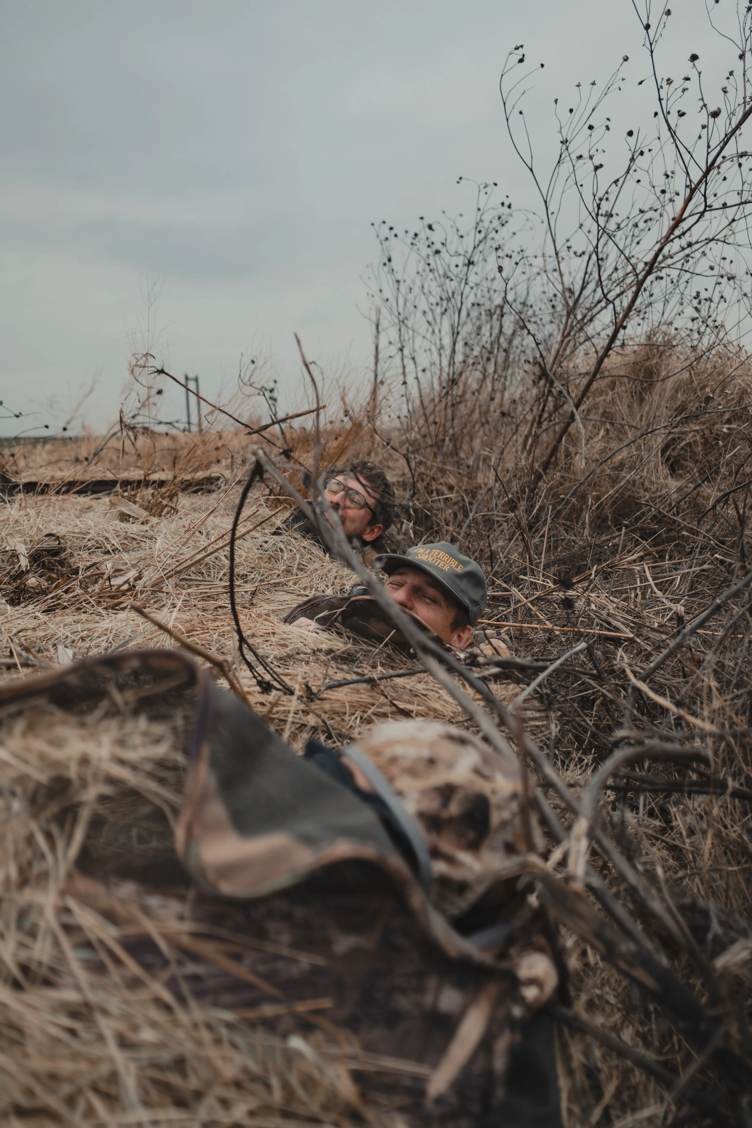 Two men lie in a trench surrounded by dry grass and branches, smiling and looking upward.