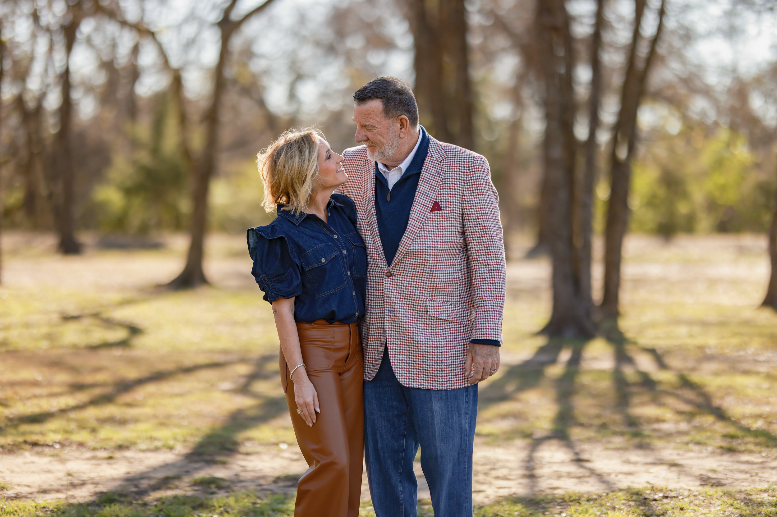 A middle-aged man and a young woman stand close together outdoors in a park, smiling at each other against a background of trees with sunlight filtering through.