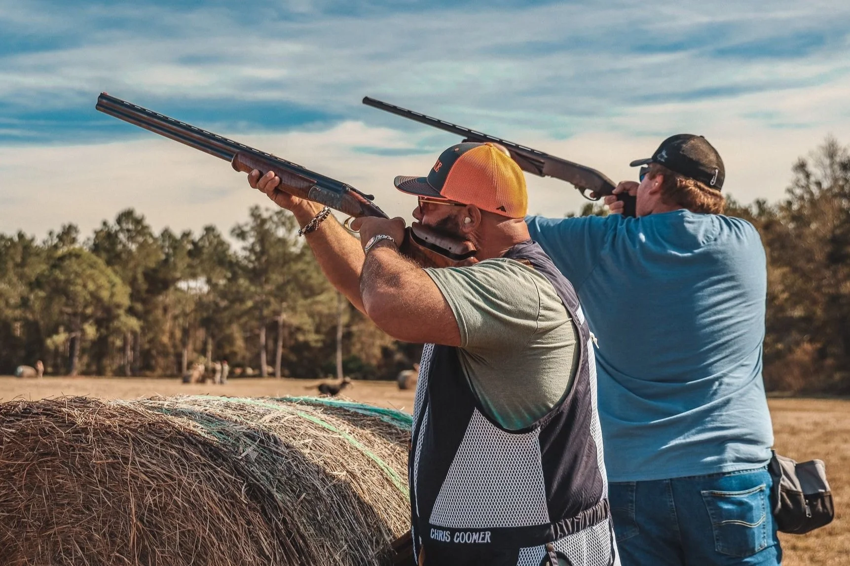 Two men shooting shotguns outdoors on a sunny day near a hay bale, with trees in the background.