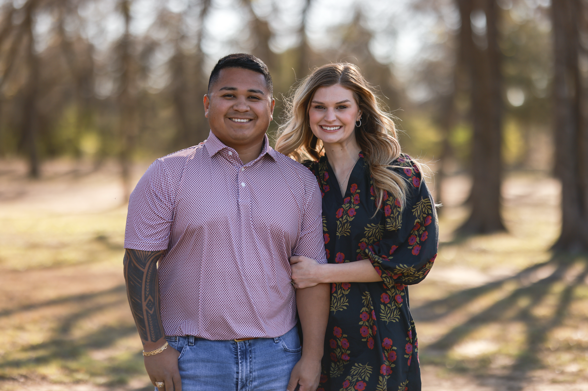 A smiling man and woman standing close together outdoors in a wooded area during daylight, with the woman holding the man's arm.