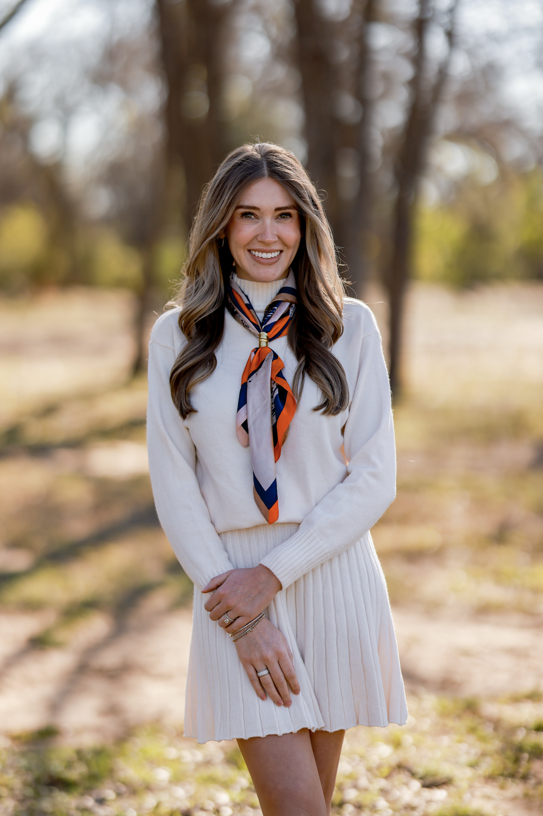 A young woman with long wavy hair smiling outdoors in a park during fall, wearing a cream sweater, a beige skirt, and a colorful scarf tied around her neck.