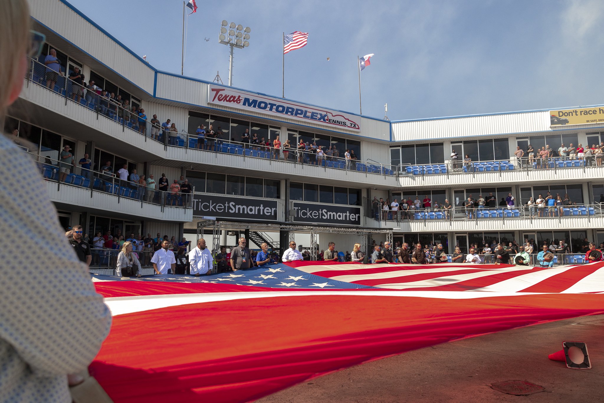 People gather around a large American flag displayed at Texas Motorplex in Ennis, Texas, during a patriotic event. The grandstands are filled with spectators, and the sky is clear with signs and flags visible.
