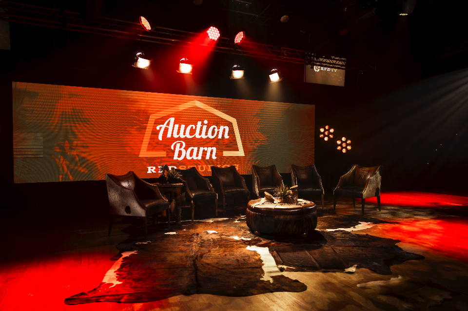 Stage setup with six armchairs arranged in a semi-circle, a central round coffee table, and a cowhide rug on the floor. Behind the chairs is a large screen displaying the logo 'Auction Barn' with red lighting overhead, indicating an auction or event 