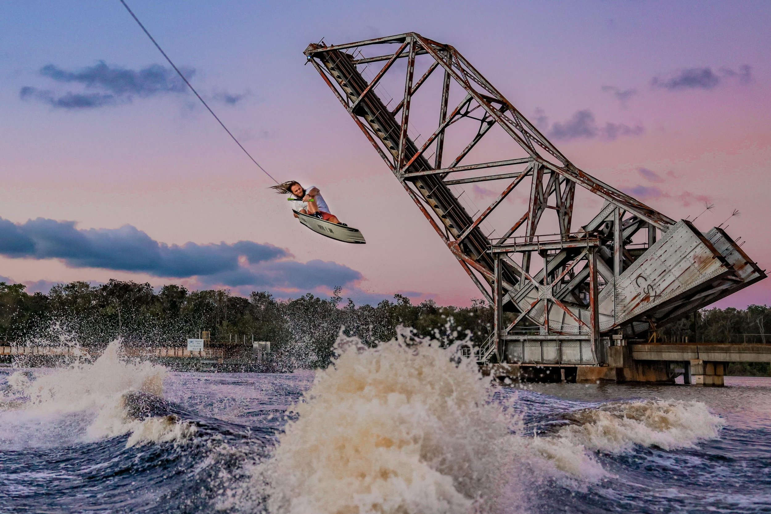 A wakeboarder airborne off the wake at sunset beneath an old railroad bridge.