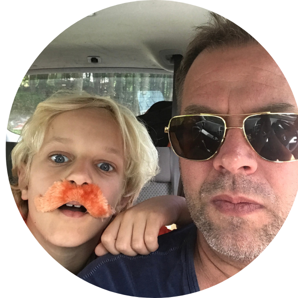 A young boy with messy blond hair wearing a fake orange mustache and a man with sunglasses inside a vehicle, with trees visible through the window.