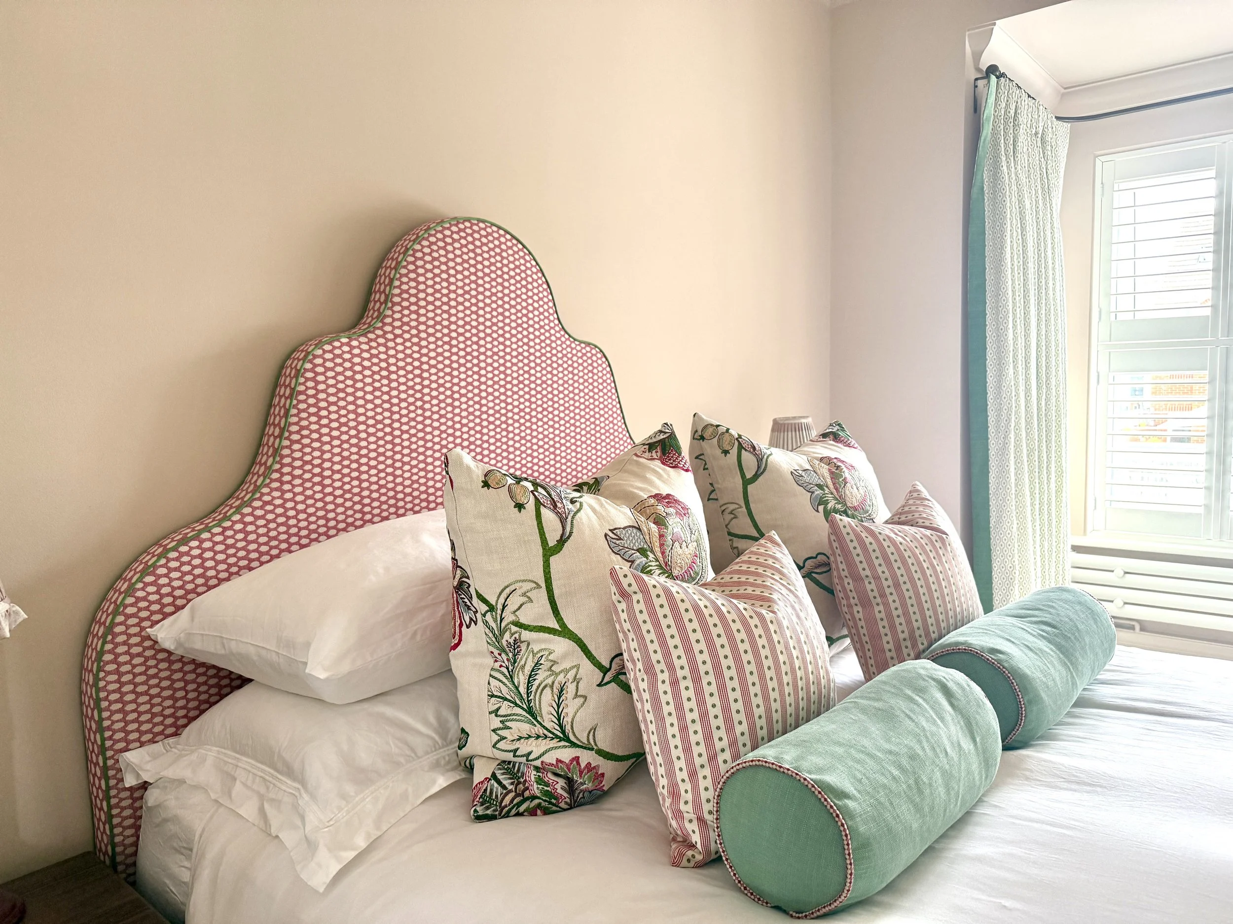 A bedroom with a bed featuring a patterned pink and white headboard, multiple pillows with floral and striped patterns, and a large green bolster pillow. A window with white shutters and patterned curtains is visible in the background.