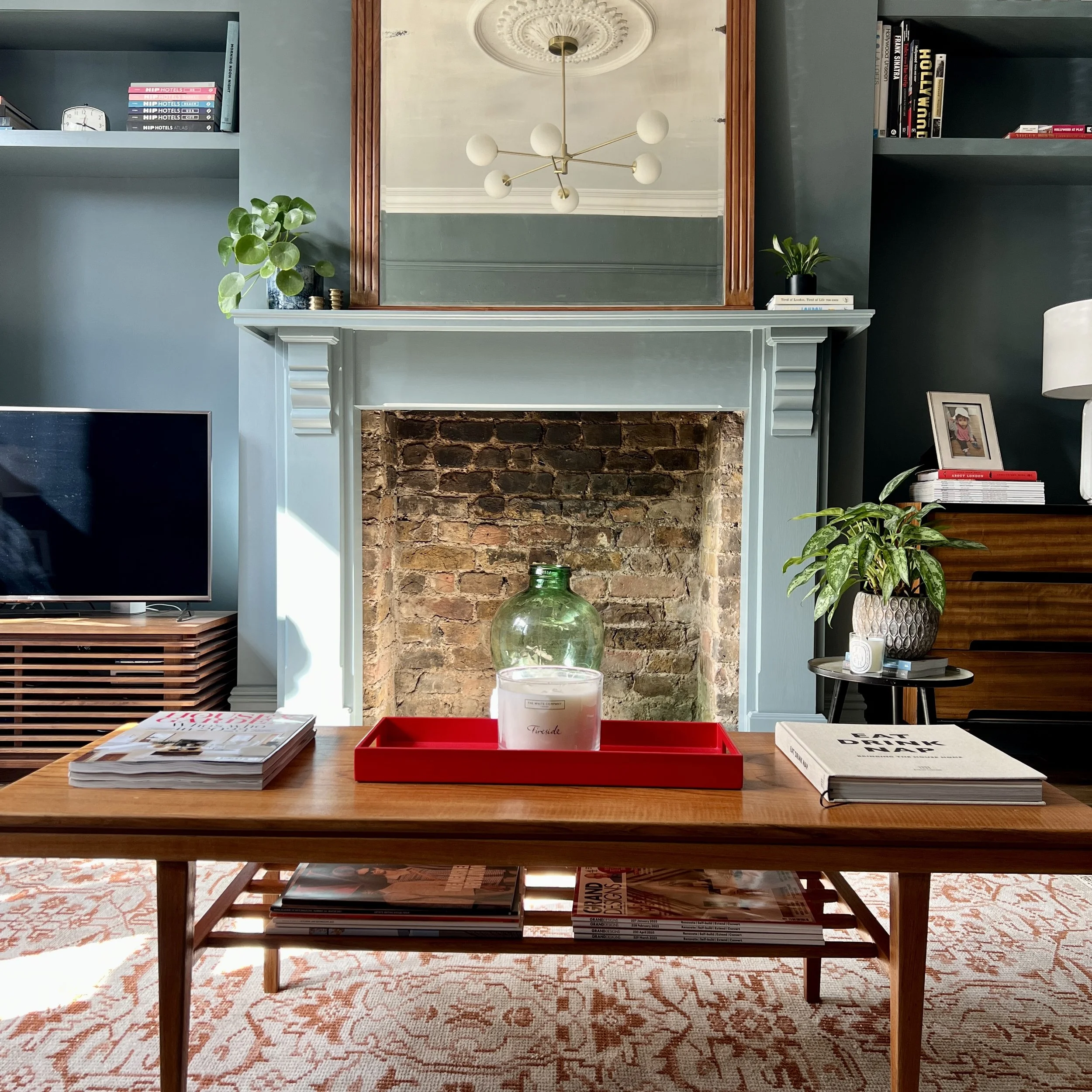Living room decor featuring a brick fireplace with a white mantel, mirror, plants, books, magazines, a candle, and a television, with a wooden coffee table on a patterned rug.