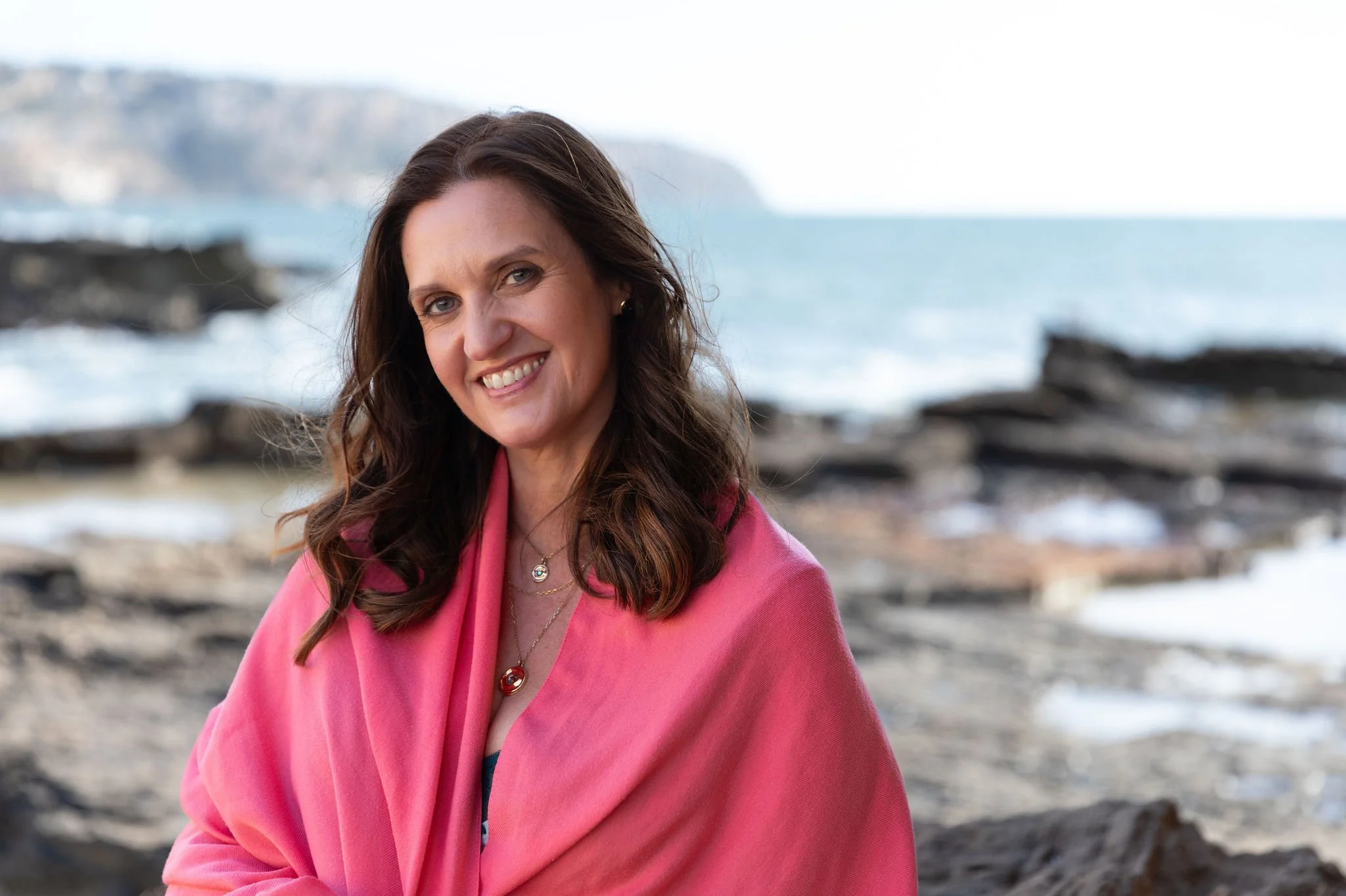 Romy Wyser smiling on a rocky beach in Mallorca, wearing a bright pink wrap with the sea behind her.