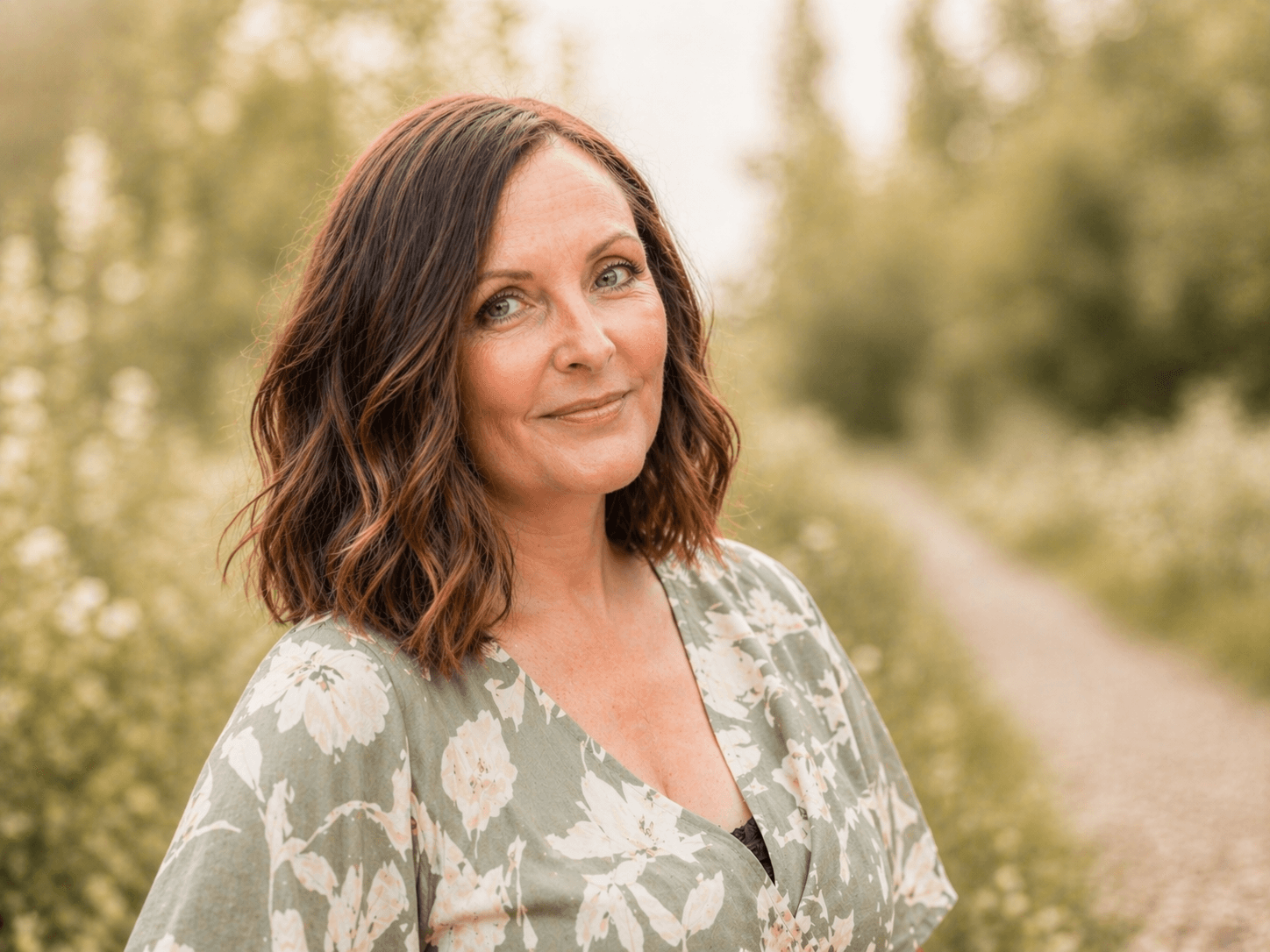Portrait of Emma Read outdoors in soft natural light, wearing a sage green floral dress and smiling beside a countryside path