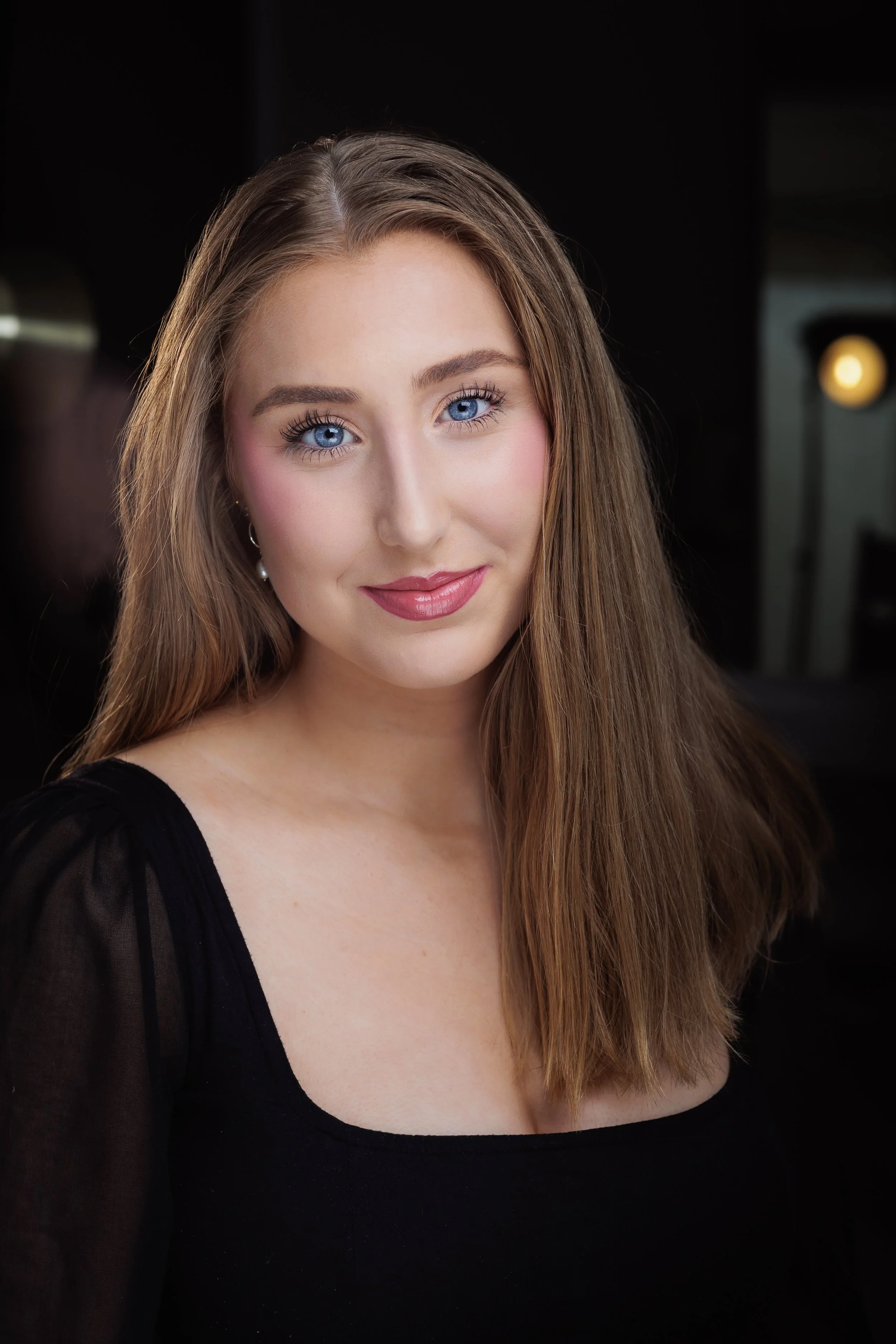 Close-up portrait of a young woman with long, light brown hair, blue eyes, wearing a black top, in a professional setting.