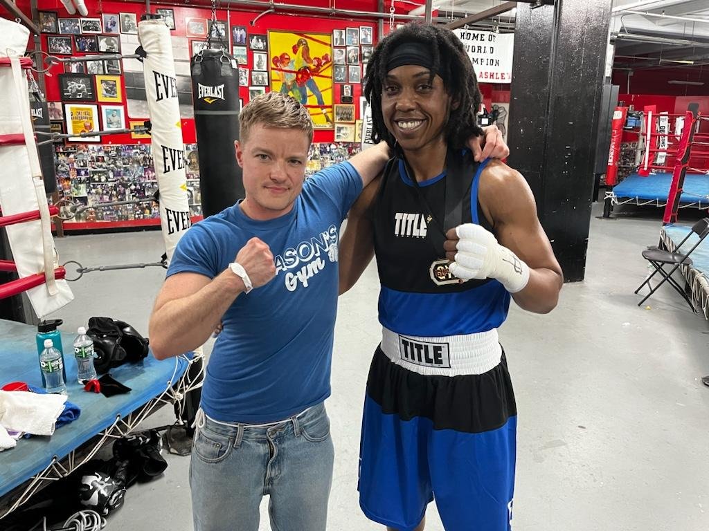 A young male and a female boxer posing together inside a boxing gym, both flexing their arms with fists clenched. The woman is wearing boxing gloves and boxing gear, while the man is dressed casually in a blue t-shirt and jeans.