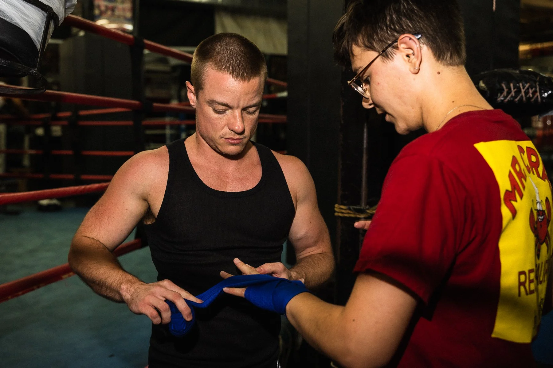 A male boxer in a black tank top having his hands wrapped by a young woman with glasses and a red shirt with yellow text in a boxing gym.