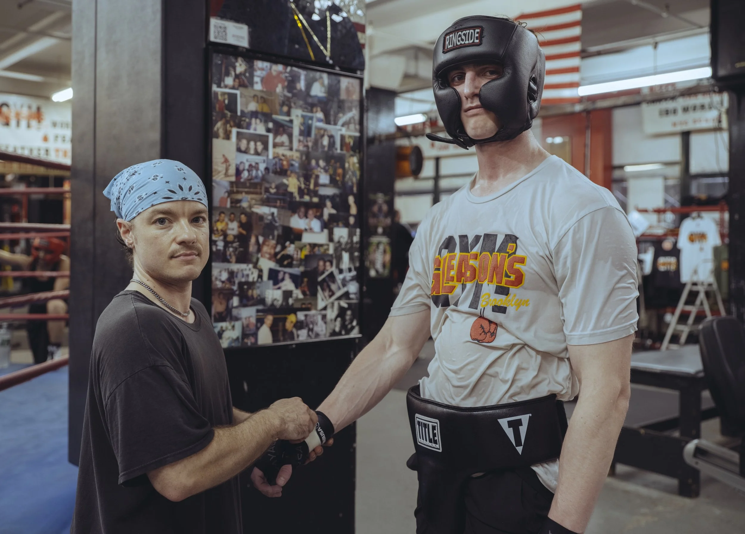 A boxer wearing a boxing helmet and gloves shaking hands with a person in a boxing gym, with photos on the wall behind them.