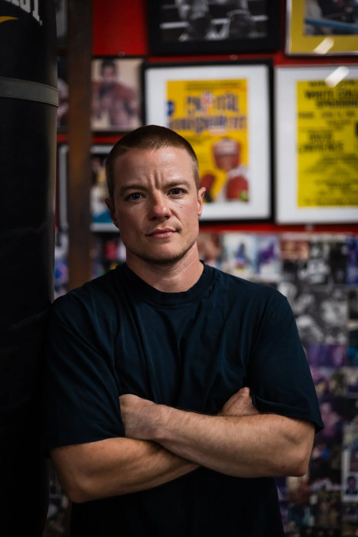 A young man with short hair and a serious expression, standing with arms crossed in a sports store. The background features posters, boxes, and shelves with sports merchandise.
