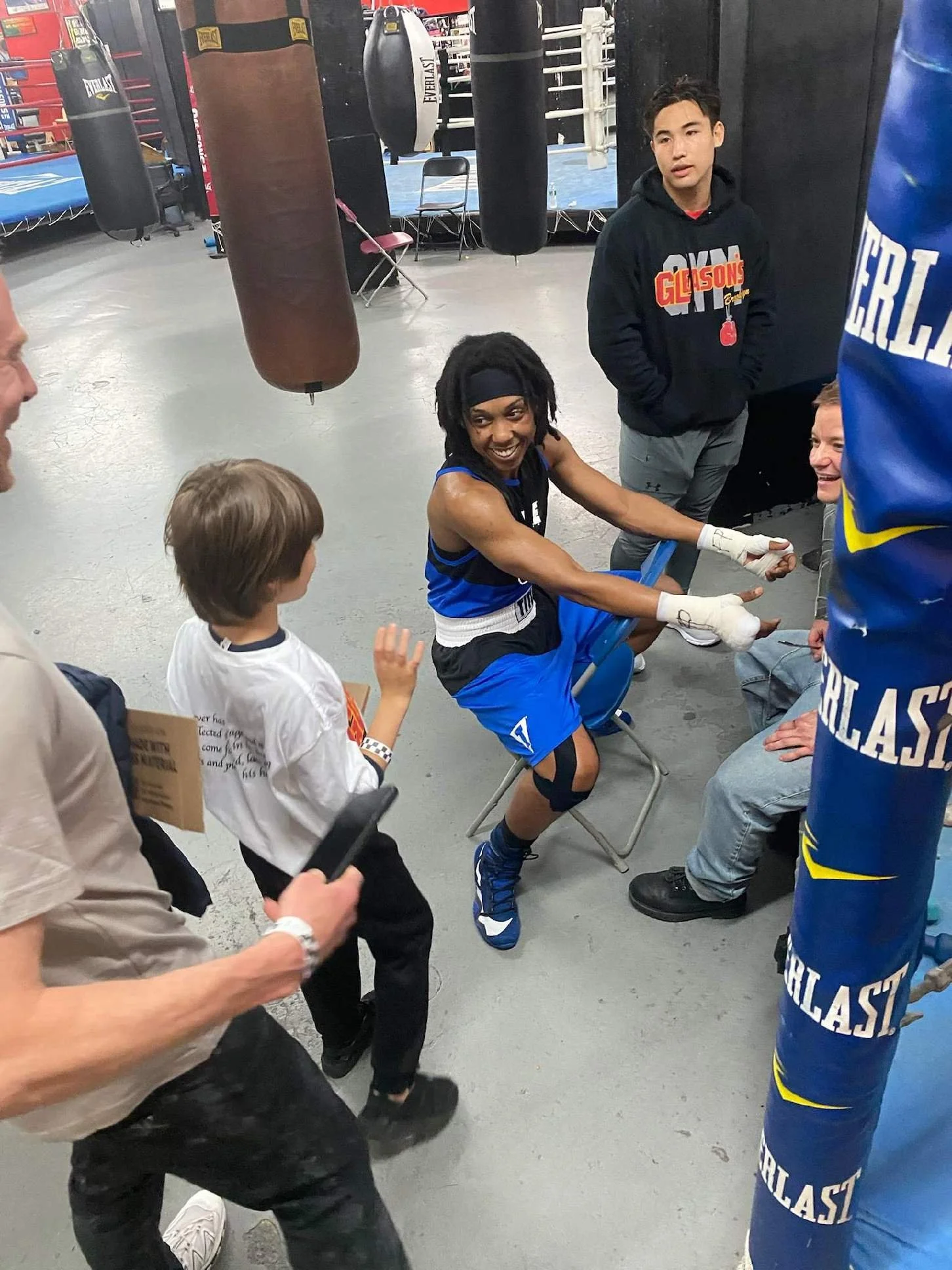 Boxers training in a gym, with one boxer sitting on a chair, wearing boxing gloves, smiling, and surrounded by trainers and friends, with punching bags in the background.