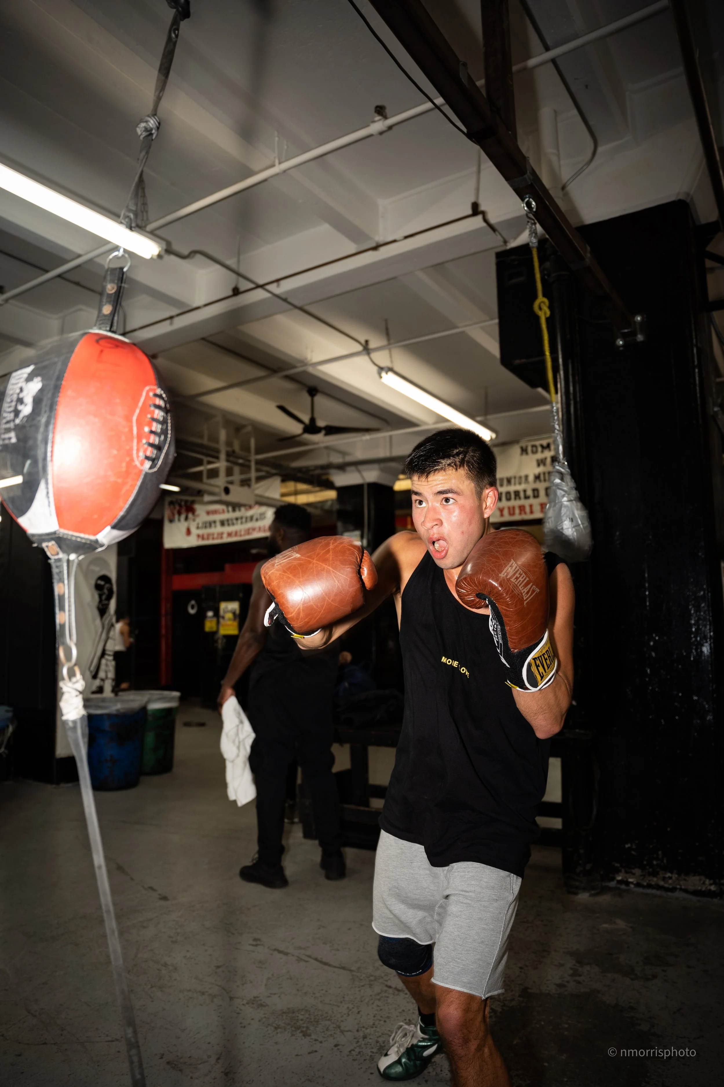 A man wearing boxing gloves and athletic shorts practicing boxing in a gym, with a punching bag hanging nearby.