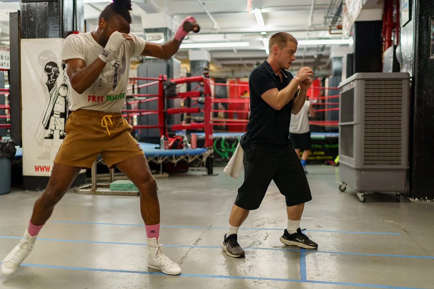 Two person practicing boxing or kickboxing in a gym. One is wearing a white t-shirt, tan shorts, and boxing gloves, while the other is in a black shirt and shorts, positioned near a padded punching bag.