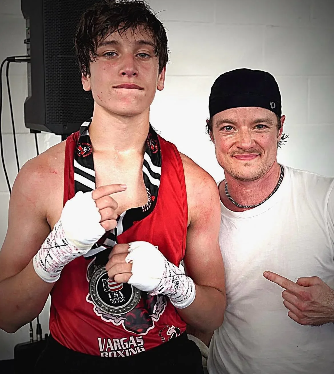 Young male boxer with boxing gloves and medal, posing with a man in a white shirt and black headband inside a gym.