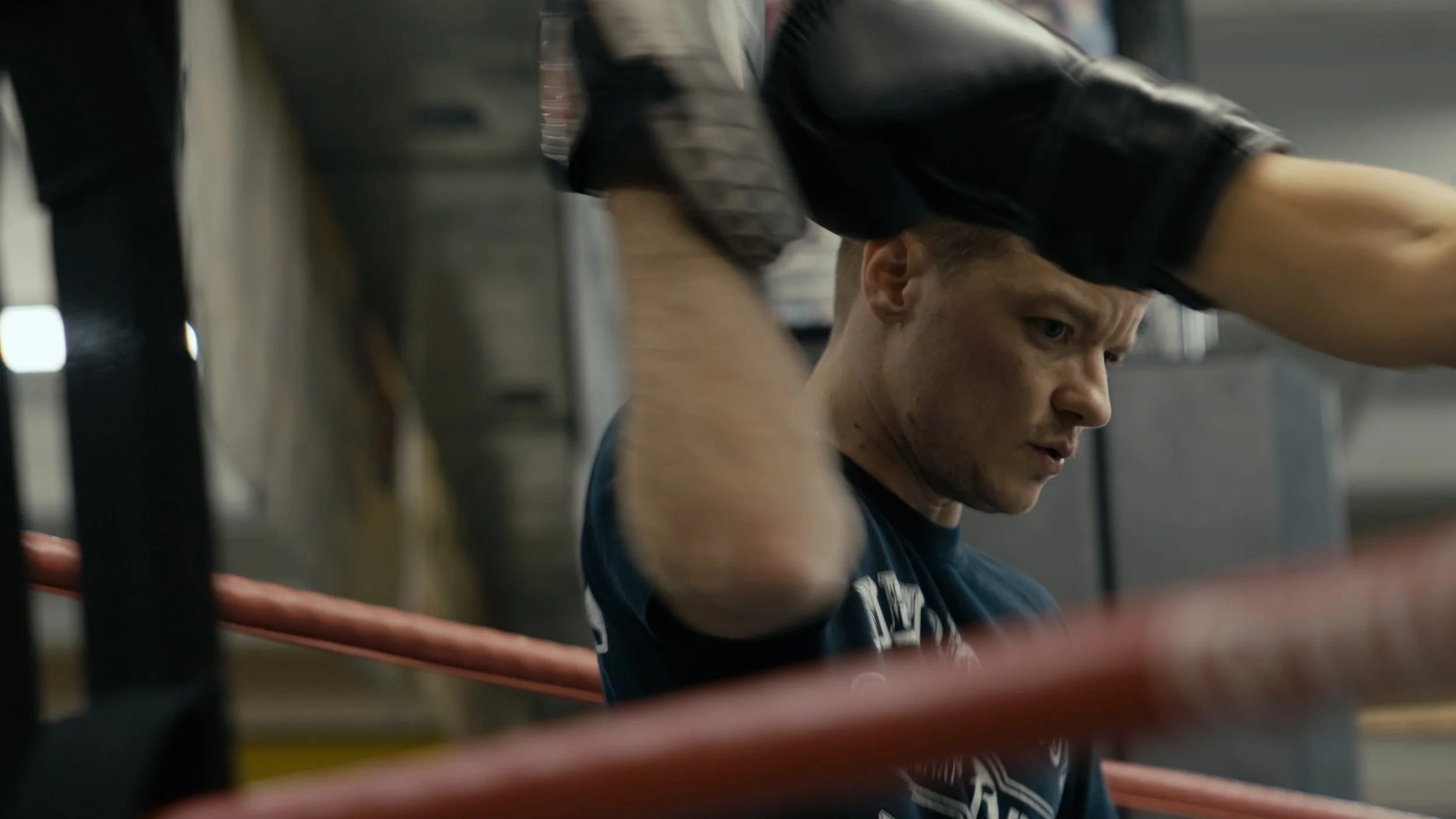A young man with short hair, wearing a dark T-shirt, sits in a gym surrounded by boxing equipment. He appears focused and slightly tired, resting against the ropes of a boxing ring.
