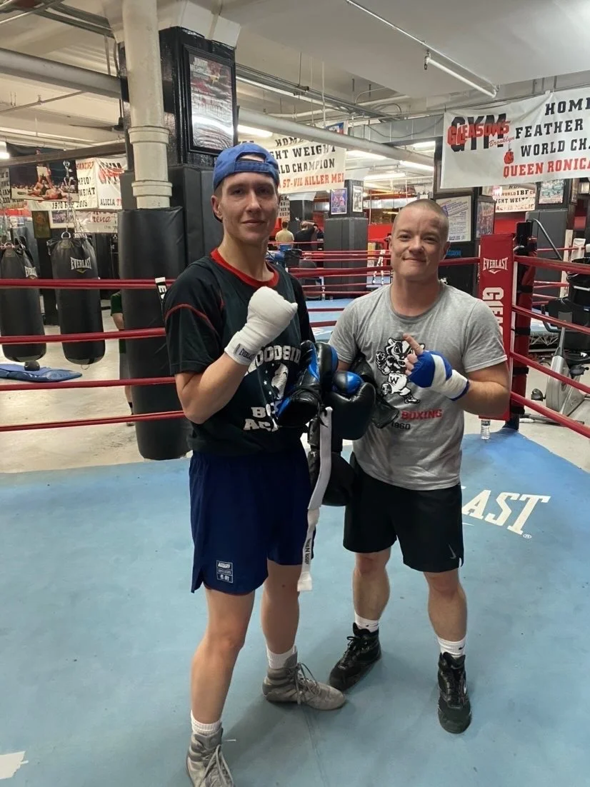 Two men in boxing gear standing in a boxing gym, posing with fists raised. The gym has punching bags and boxing posters in the background.