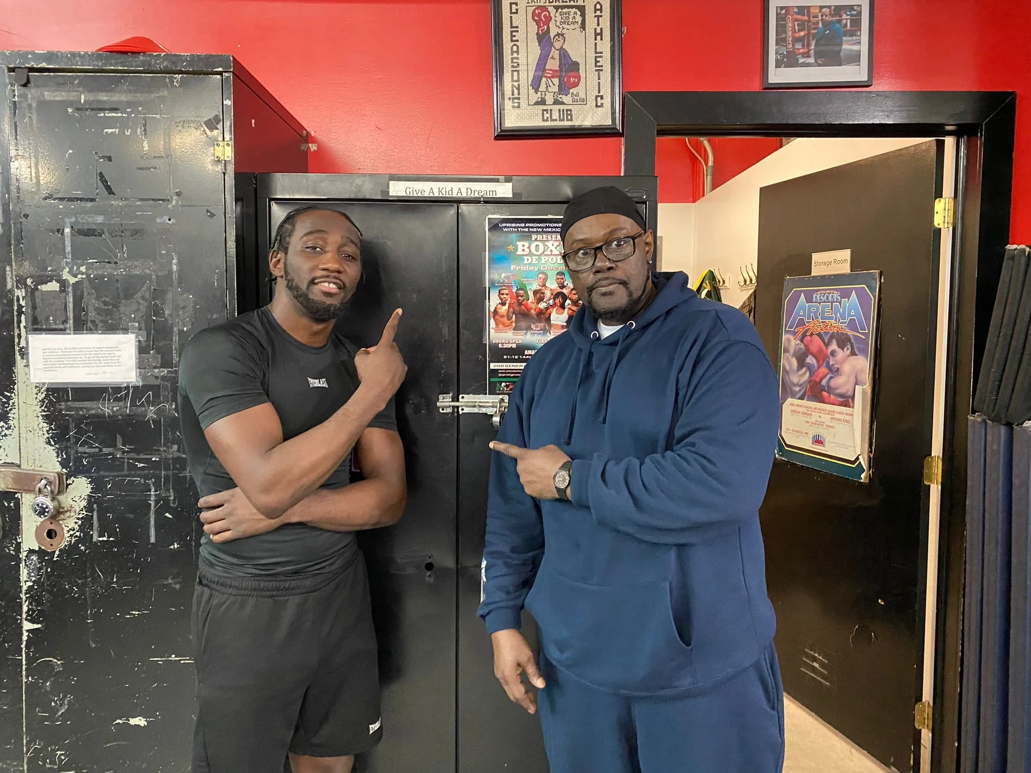 Two men standing inside a gym near a black locker. The man on the left has braided hair and a beard, wearing a black athletic shirt and shorts, pointing at the locker. The man on the right is wearing glasses, a black cap, and a blue hoodie, pointing 