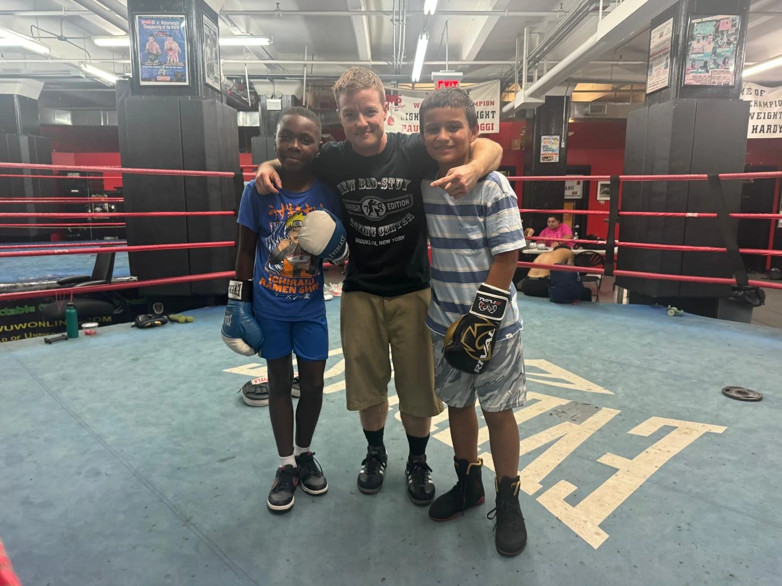Two young boys wearing boxing gloves and shorts stand with a coach in a boxing gym. The coach has his arms around them, and they are all smiling. The gym has boxing rings, equipment, and posters in the background.