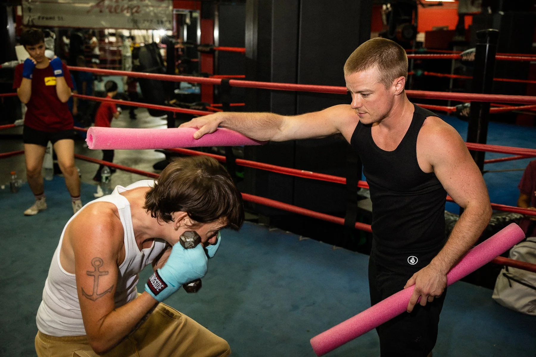 A person in boxing gloves and a white tank top practices boxing in a gym, while a man in a black tank top holds pink foam rollers above her. In the background, other people are training, including a young boy.
