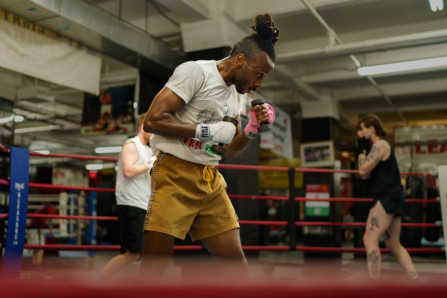 A man with a top knot hairstyle and a beard, wearing a white t-shirt and yellow shorts, is standing in a boxing ring with red ropes and holding pink and black boxing gloves, practicing boxing. Two women are in the background, one with tattoos, wearing black and white, and another with a towel around her waist.