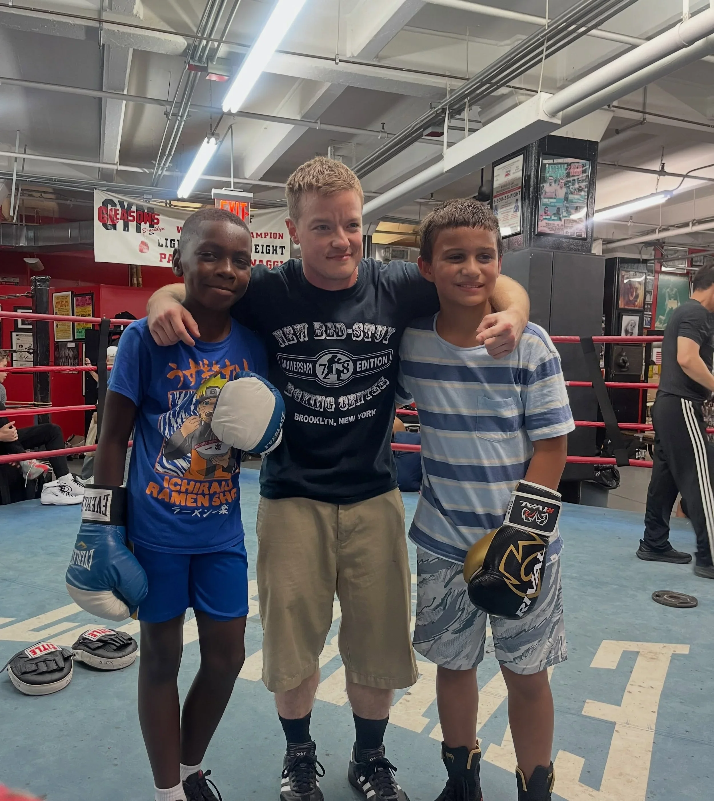 Three young males in boxing gear standing in a gym with boxing ring ropes around them. Two of them are wearing boxing gloves, and one is holding boxing pads. They are smiling, with arms around each other, inside a gym with boxing posters and equipmen