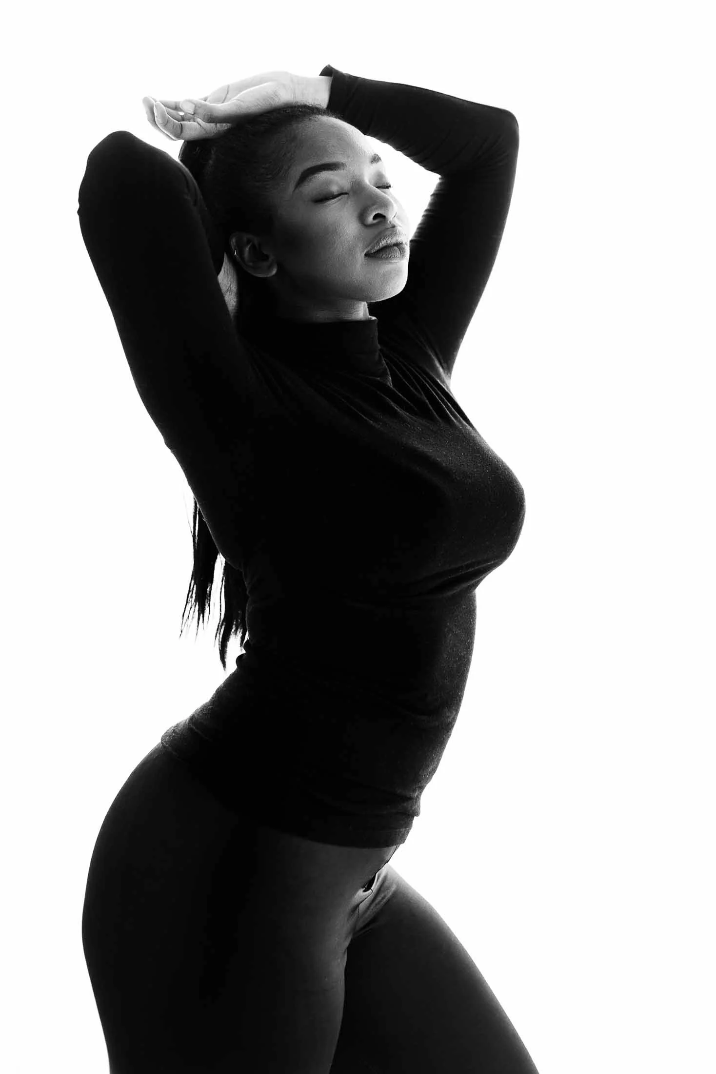Woman in long-sleeve top and pants, hands behind head. Empowered and composed modern studio image.