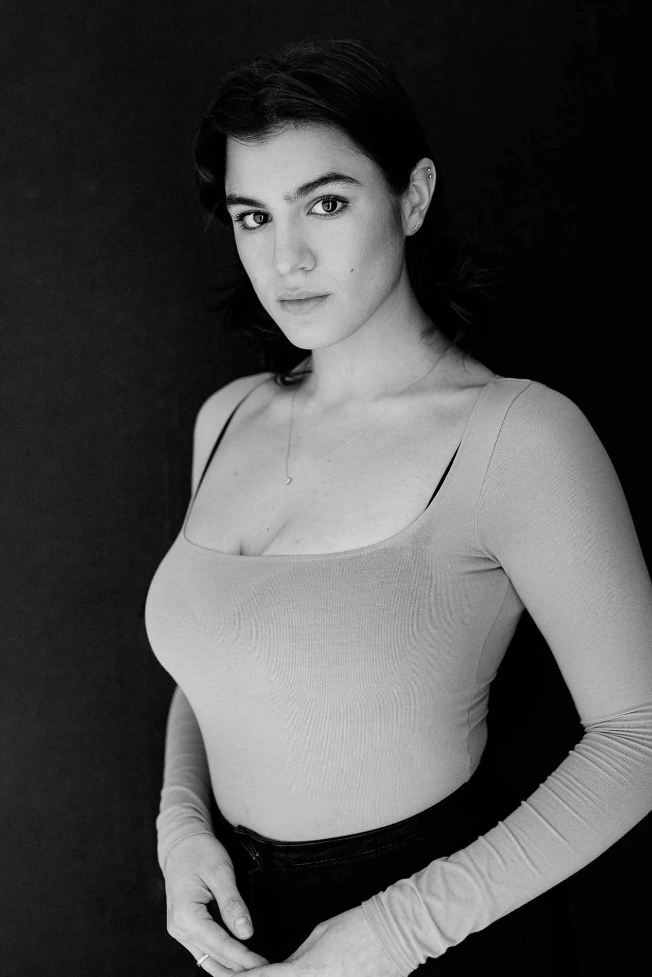 Woman with necklace and long-sleeve top standing against dark backdrop. Composed and direct black and white branding portrait.