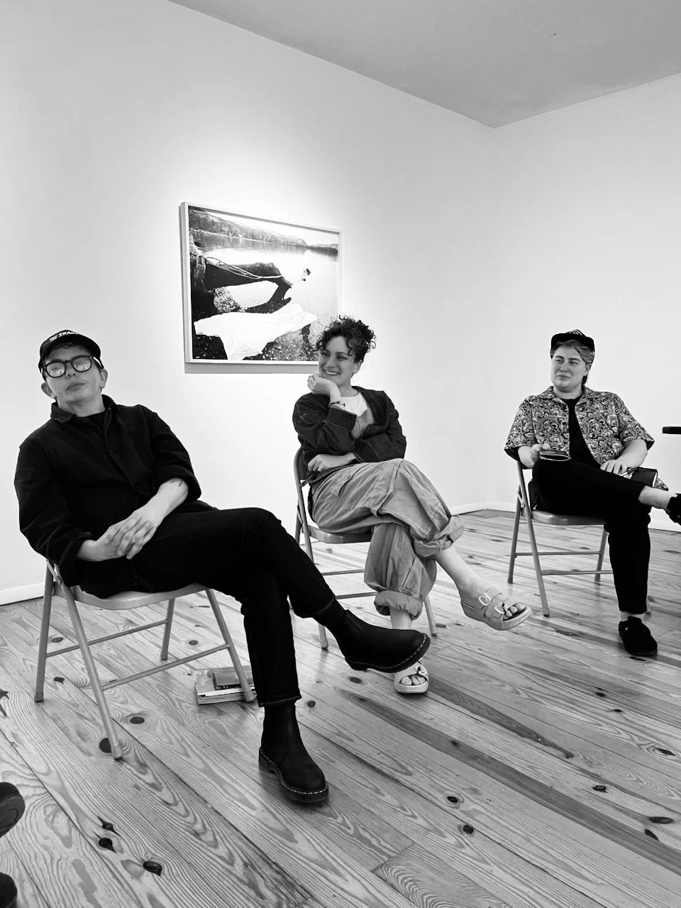 Three women sitting on chairs in an art gallery, engaging in conversation, with a framed painting of a boat on a river on the white wall behind them.