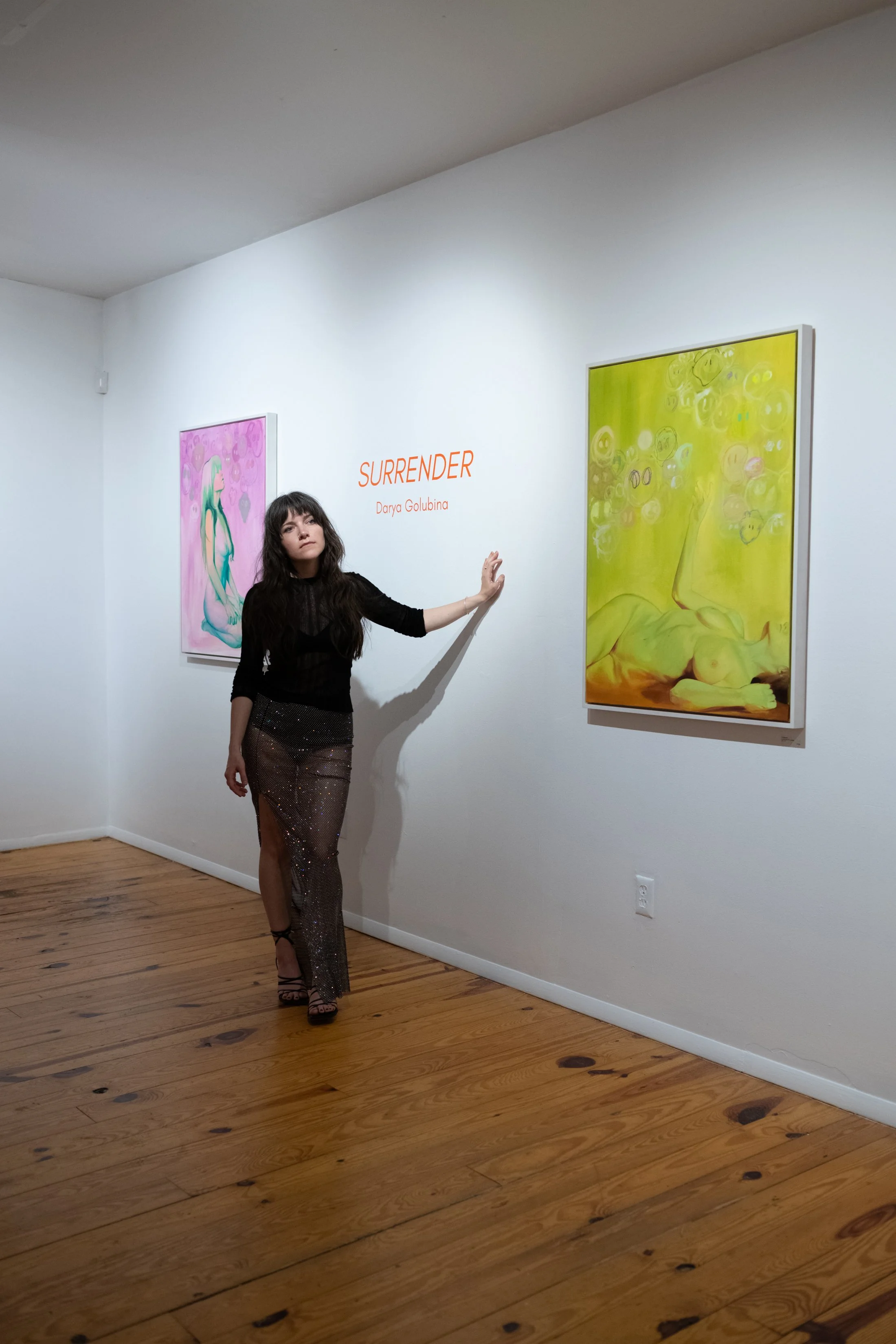 A woman in a black top and shiny black skirt stands in an art gallery next to two colorful paintings, with the word 'SURRENDER' and the artist's name, Darya Golubina, written on the wall.