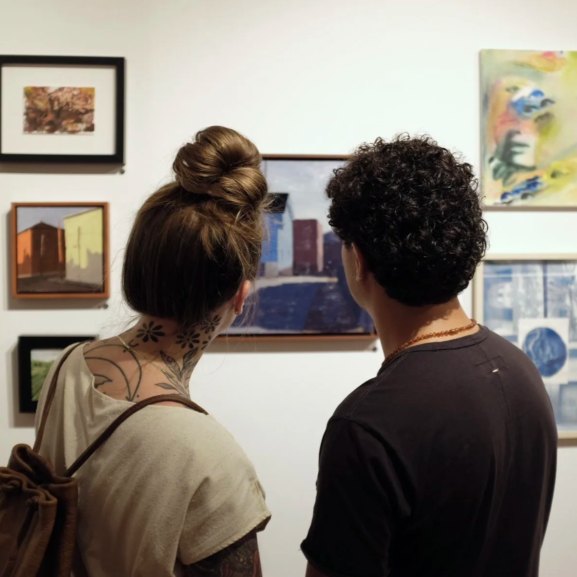 Two people viewing art in a gallery, looking at paintings and photographs on the wall.