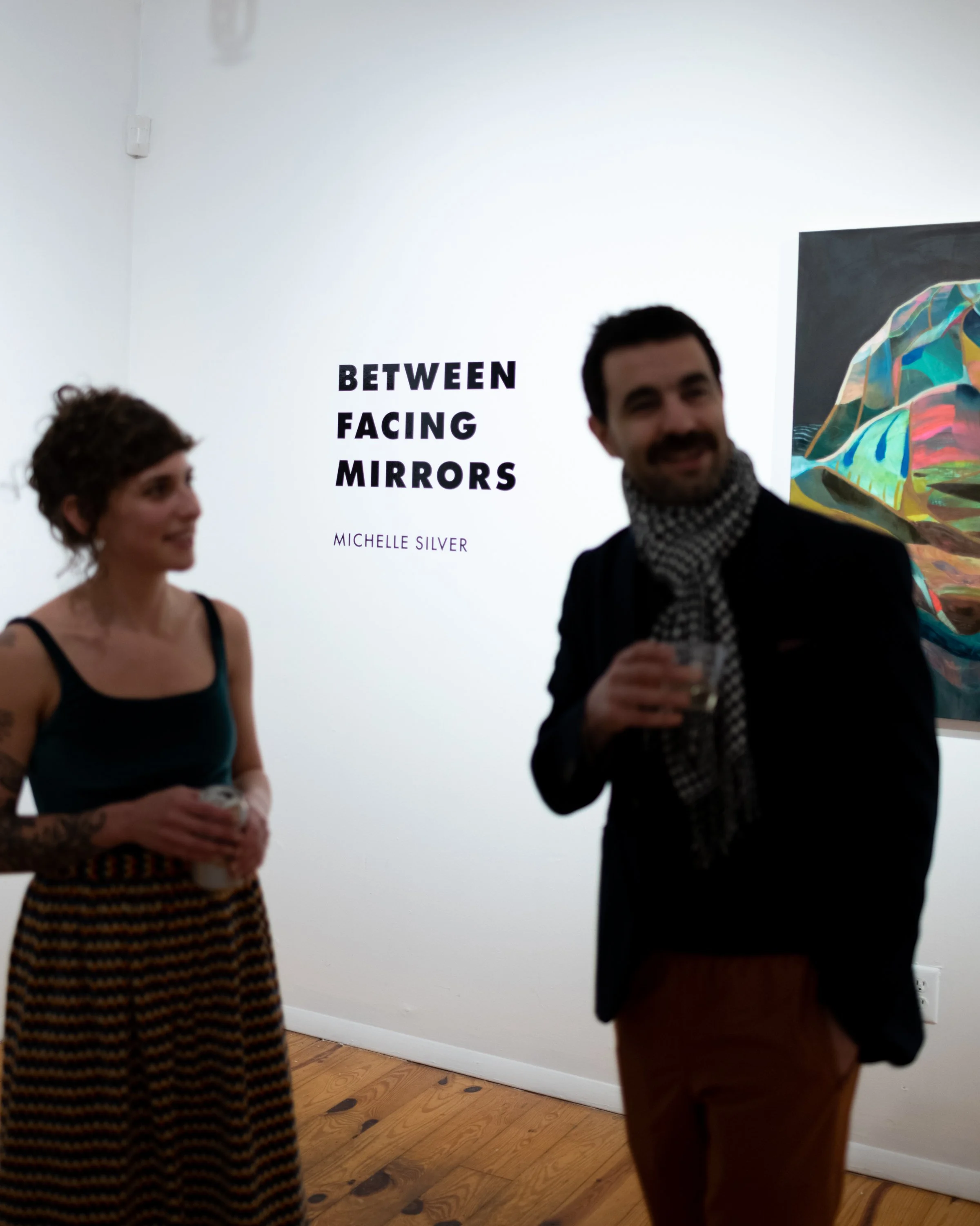 Two people standing in an art gallery, smiling and holding drinks, with a wall sign reading 'Between Facing Mirrors' by Michelle Silver in the background.