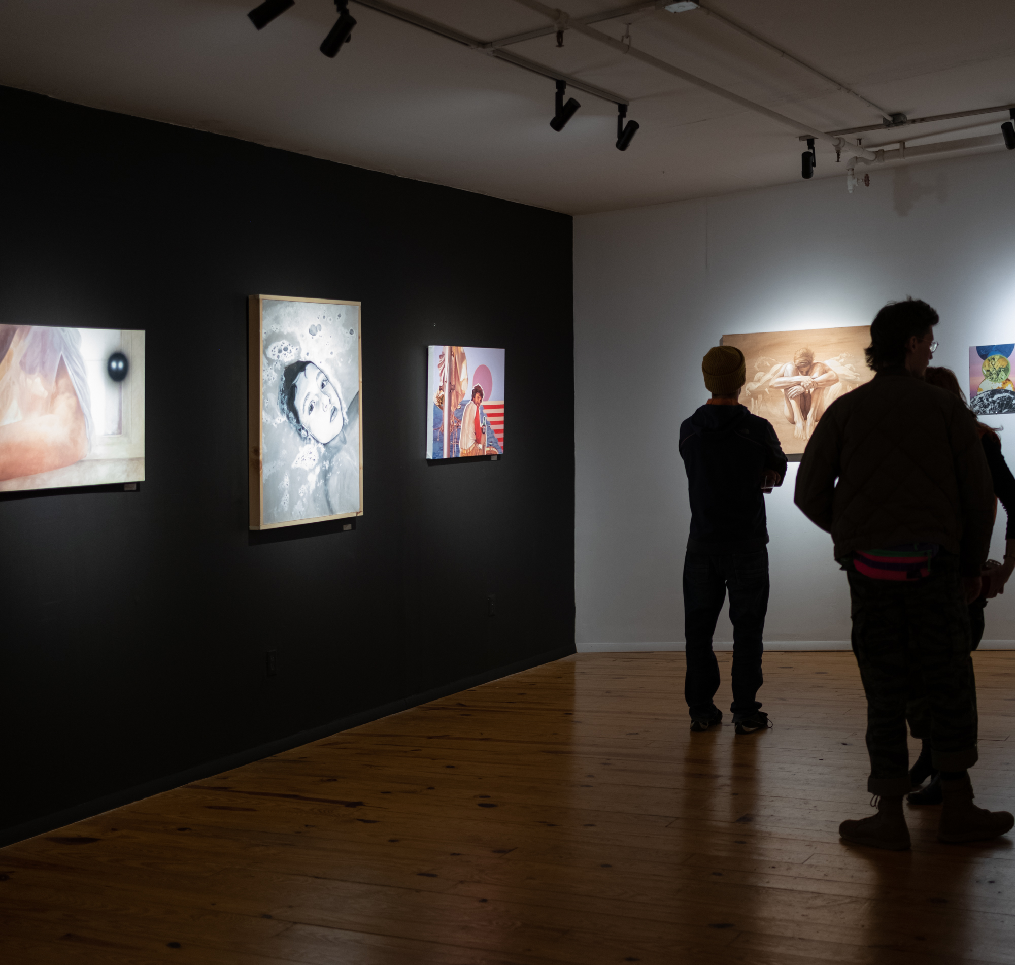 People viewing artwork in an art gallery with dark and white walls and hardwood floors.