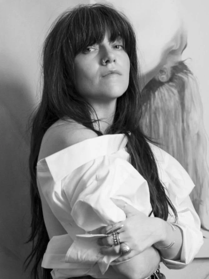 Black and white photo of a woman with long dark hair, off-the-shoulder shirt, and multiple rings, standing with her arms crossed.