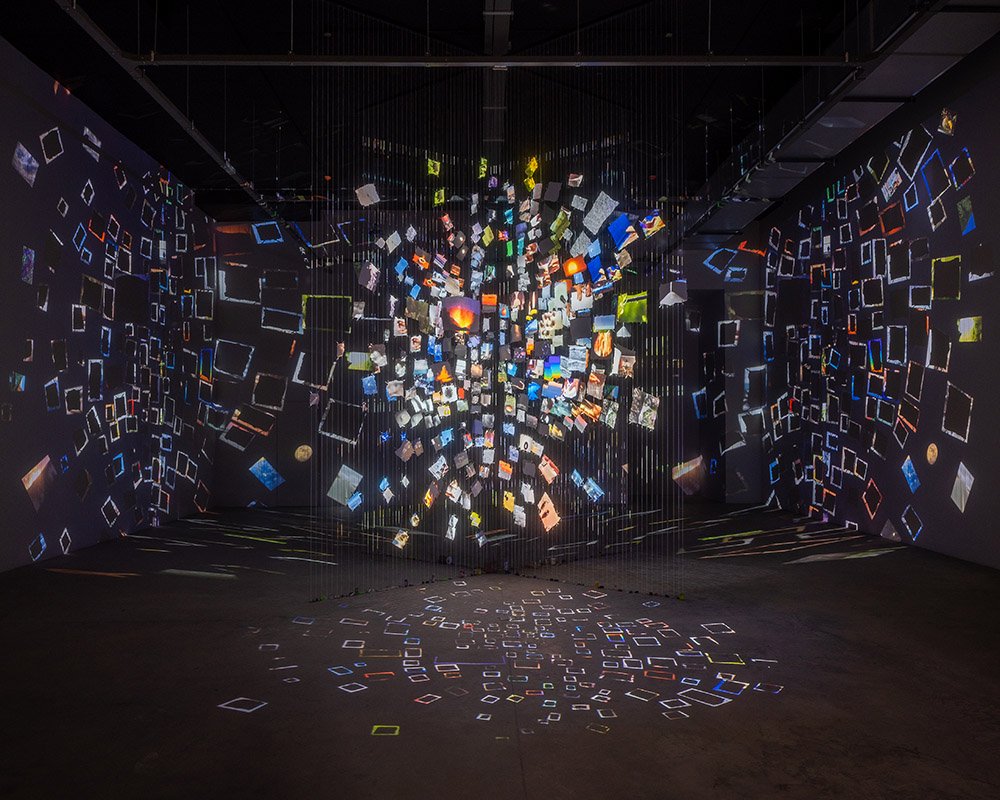 An art installation featuring numerous small, colorful, illuminated cubes and rectangles suspended in mid-air in a dark room, creating reflections and shadows on the floor and walls.