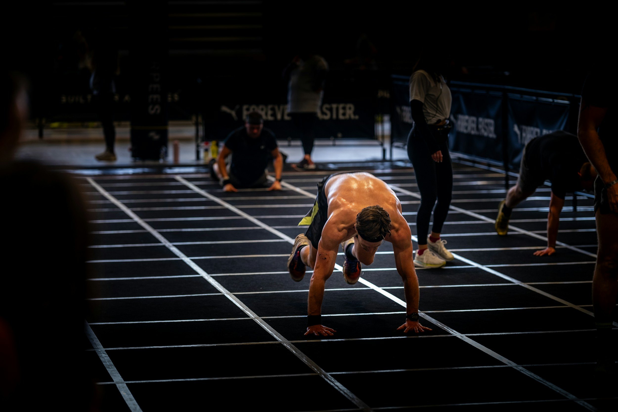 A shirtless male athlete performing the burpee part of a burpee broad jump in a lane marked with white lines on a black floor of what looks like a HYROX race or similar.