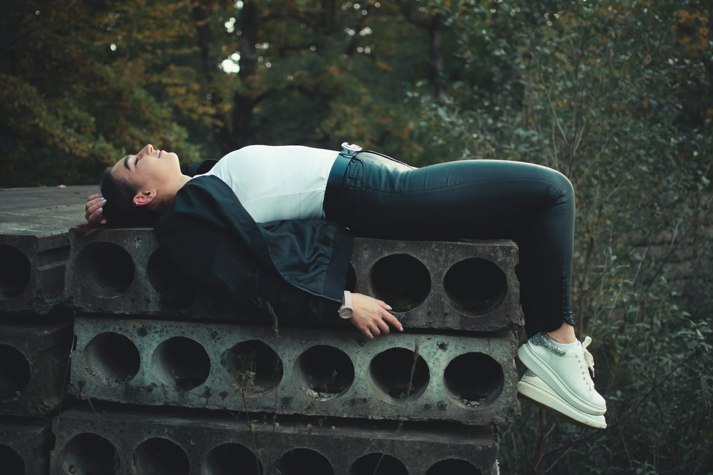 A burntout female athlete lays on her back over a stack of long concrete slabs with a forest of trees in the background