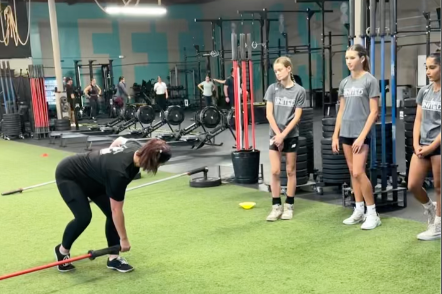 Female Coach demonstrates a hinge exercise with a barbell in a landmine set up to three young female soccer players