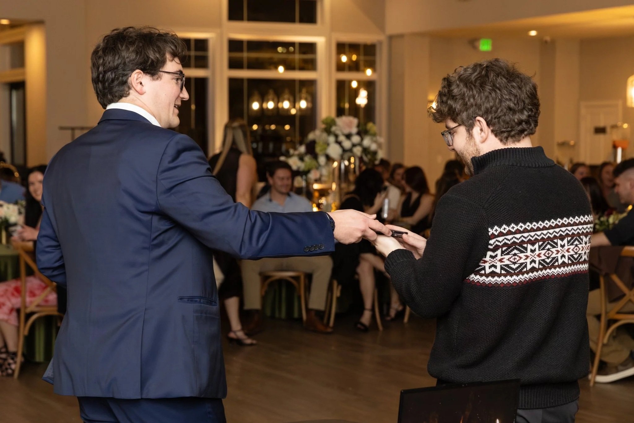 A young man wearing glasses, a suit, and a decorative bolo tie is tossing a playing card in the air while standing at a microphone in front of a wood-paneled wall. He has a name tag that reads "Germ".