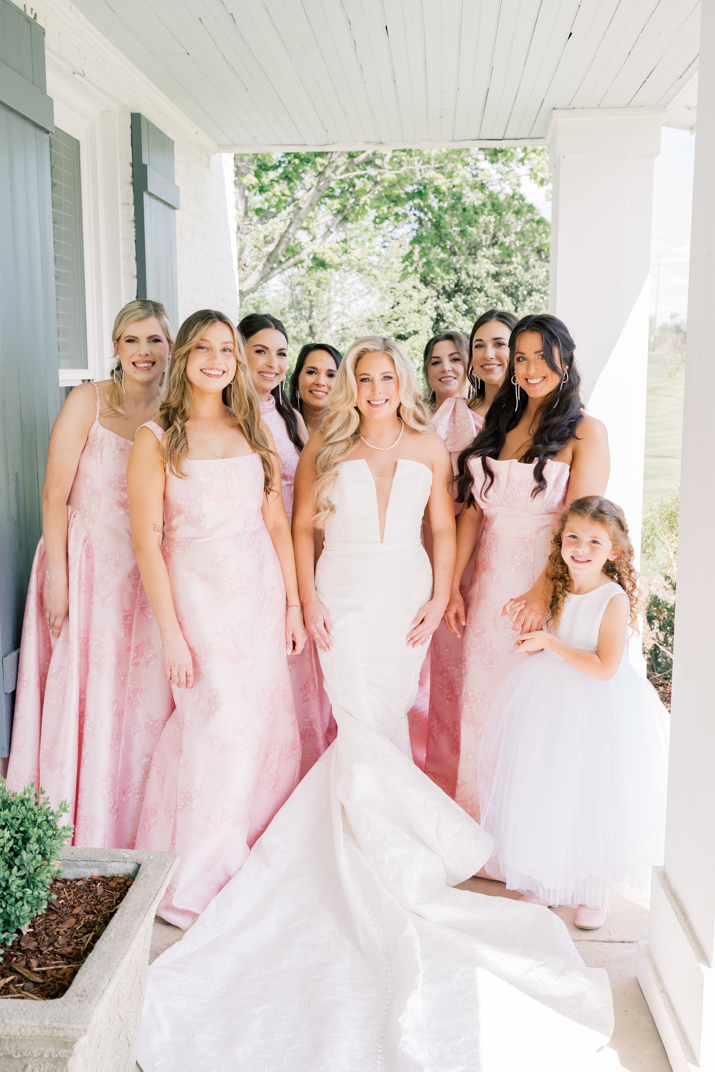 A bride in a white wedding gown with blonde hair standing with eight bridesmaids and a young girl, all smiling, on a porch with greenery in the background.