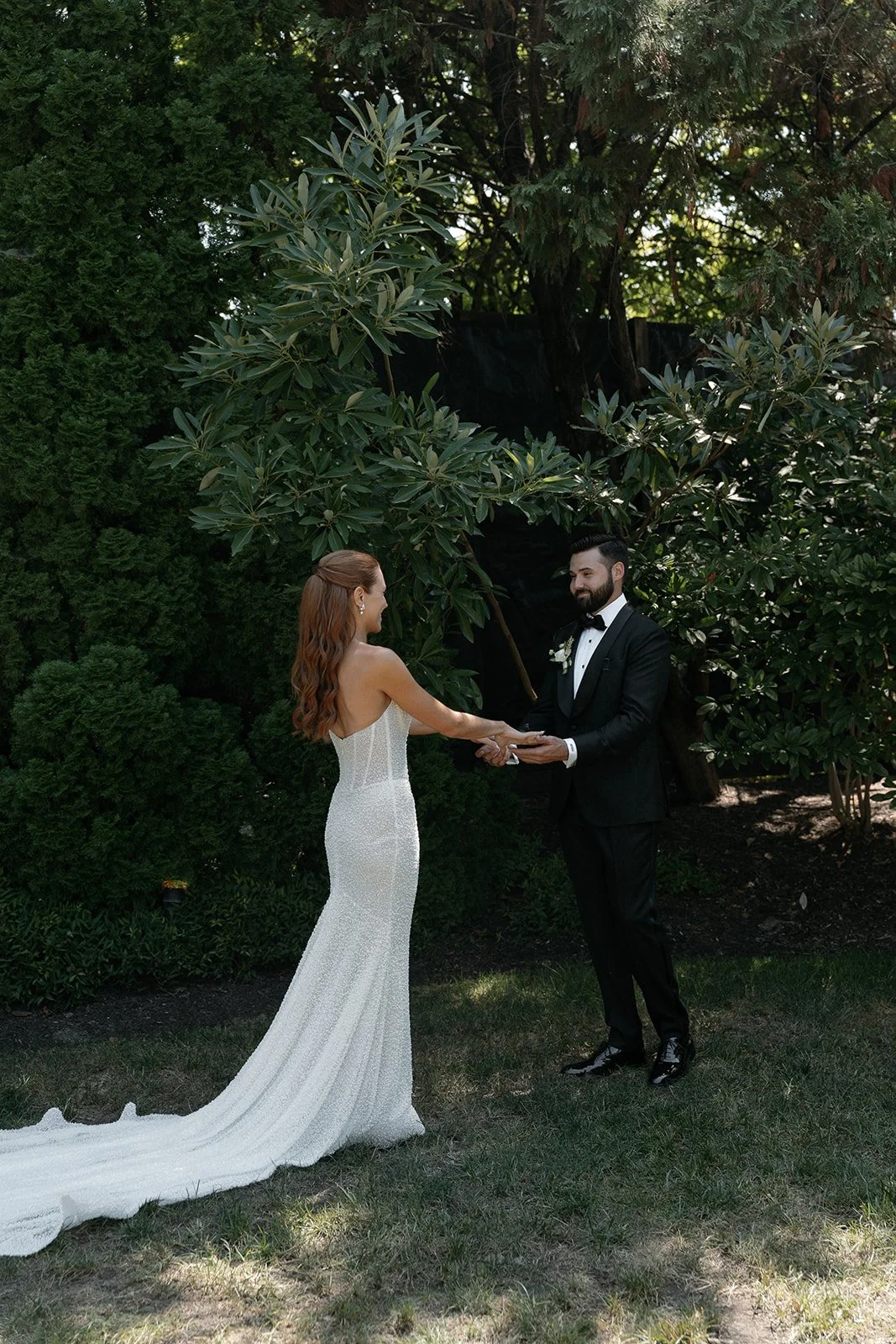 A bride and groom holding hands in an outdoor wedding ceremony surrounded by greenery, with the bride in a white, fitted gown and the groom in a black tuxedo.
