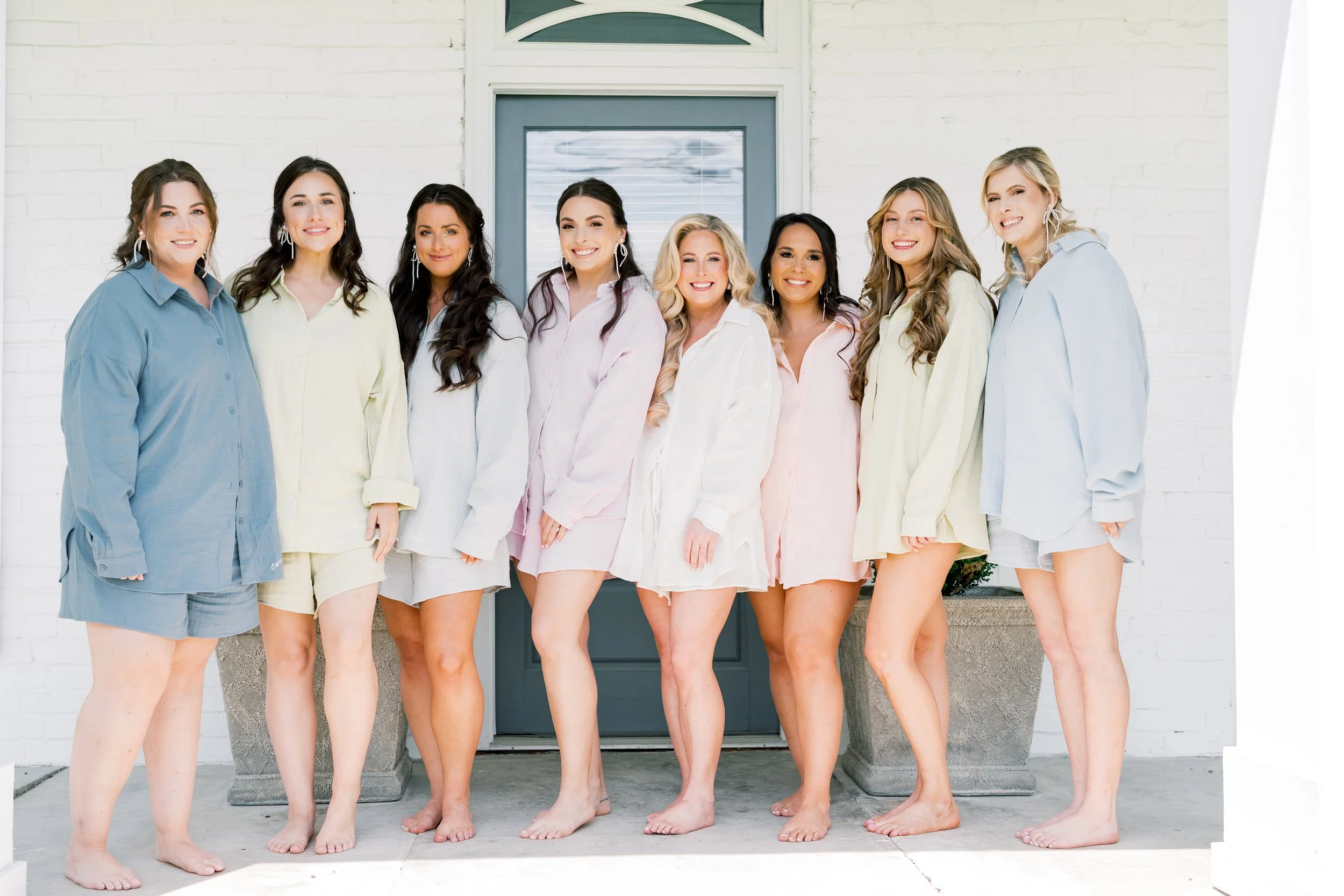 Group of eight women standing barefoot in pastel-colored pajamas outside in front of a white brick house with a blue door.