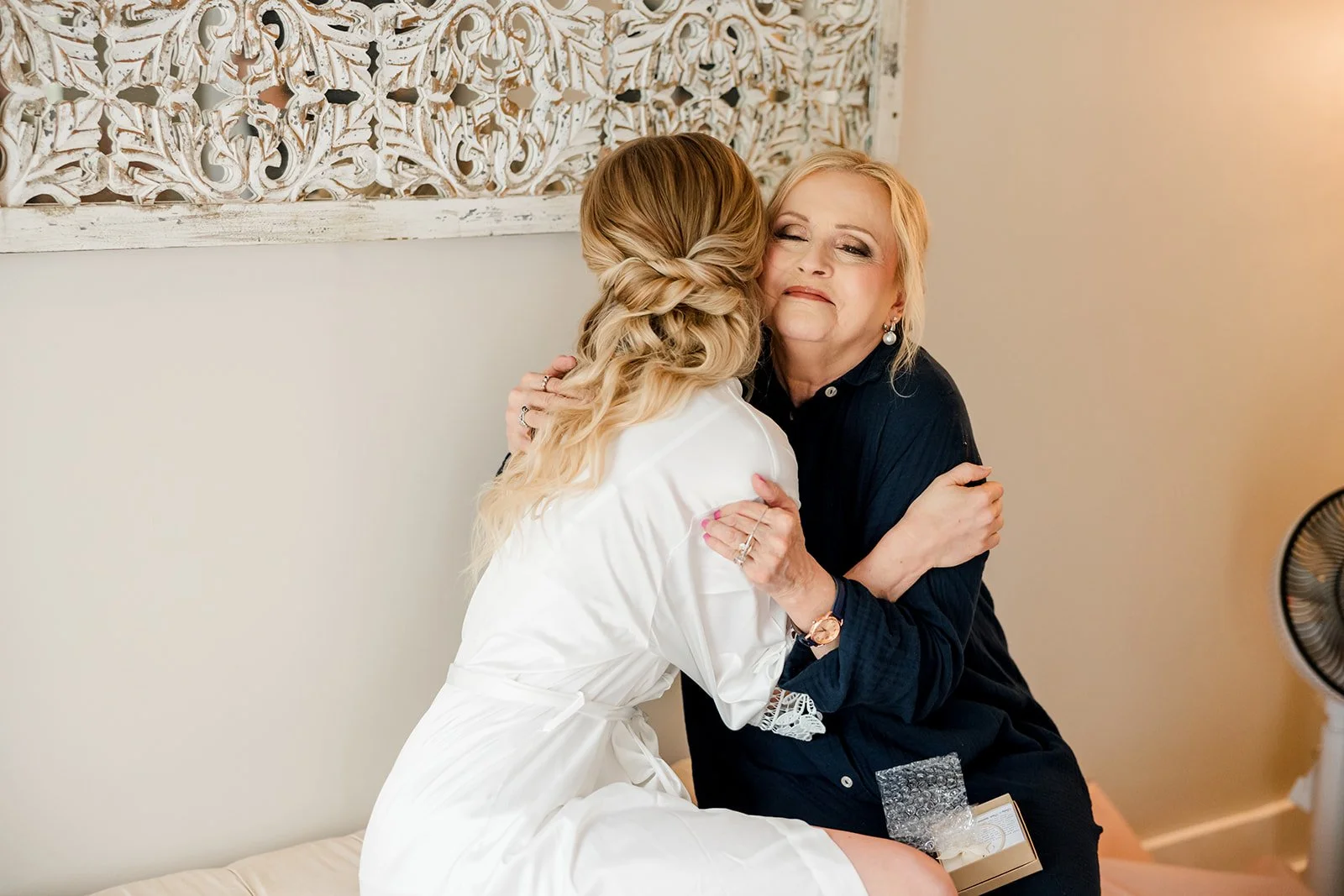 A woman in a white satin robe hugging a woman in a black shirt, both sitting on a bed in a warmly lit room, with a decorative wooden panel hanging on the wall behind them.