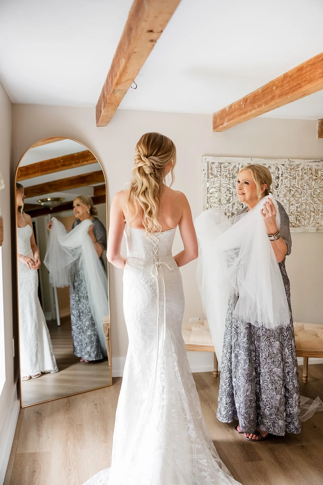 A bride in a white wedding gown standing in front of a mirror, smiling and holding the edge of her veil, as an older woman helps her with the veil in a well-lit room with wooden beams.