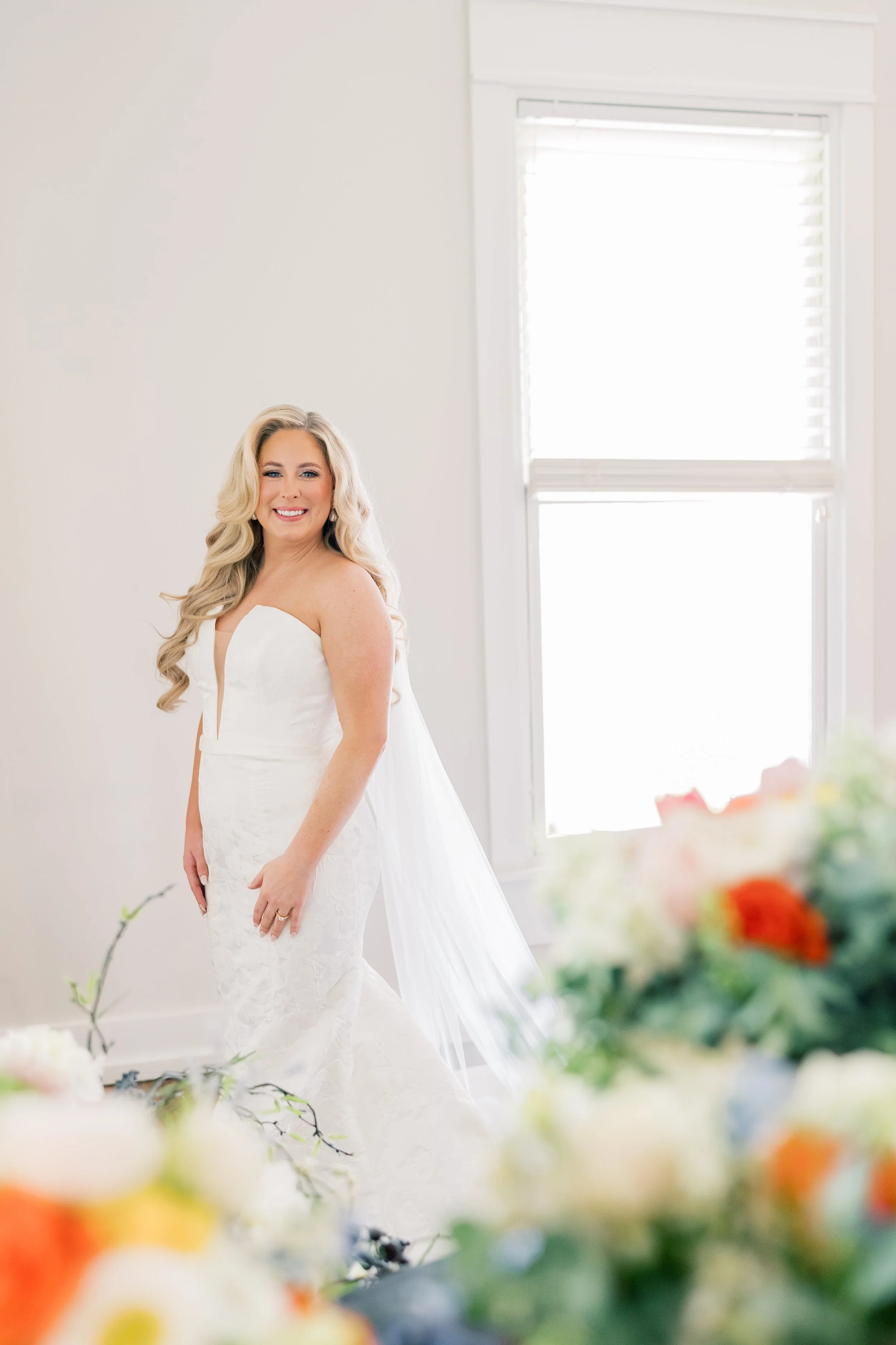 A smiling bride in a white strapless wedding gown with a sheer veil, standing in a bright room with a large window, surrounded by colorful flowers.