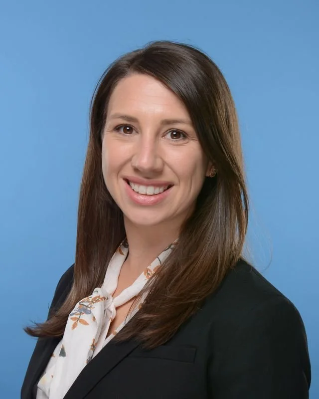 professional woman with brown hair wearing a black blazer and a white scarf with floral pattern, smiling against a blue background